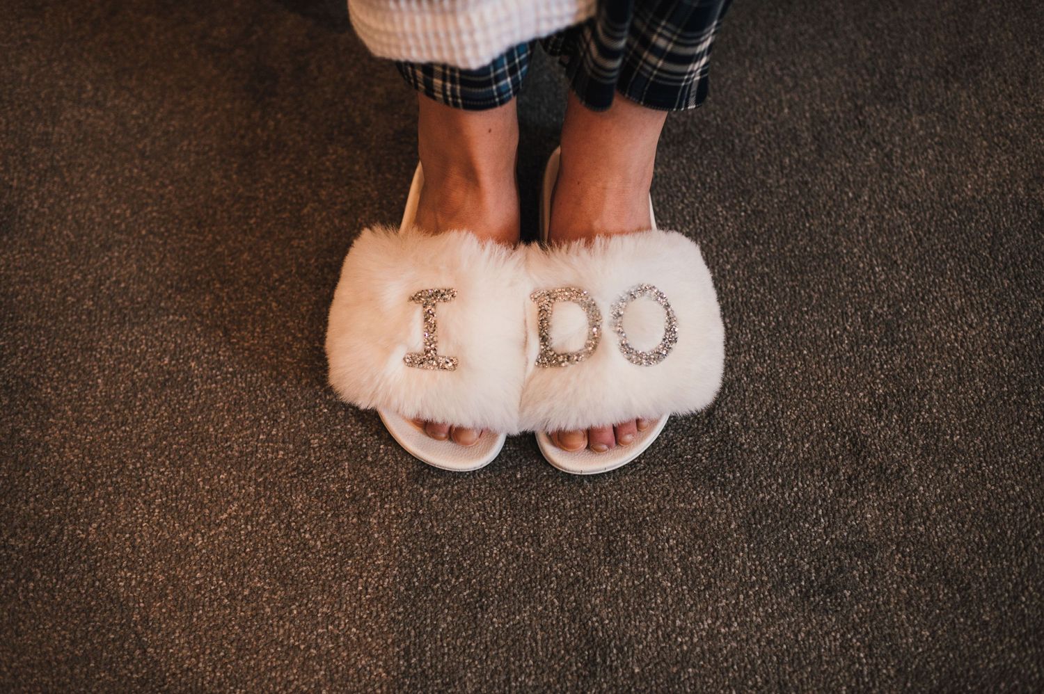 Fuzzy white slippers with 'I DO' text embroidered on them on dark carpet.