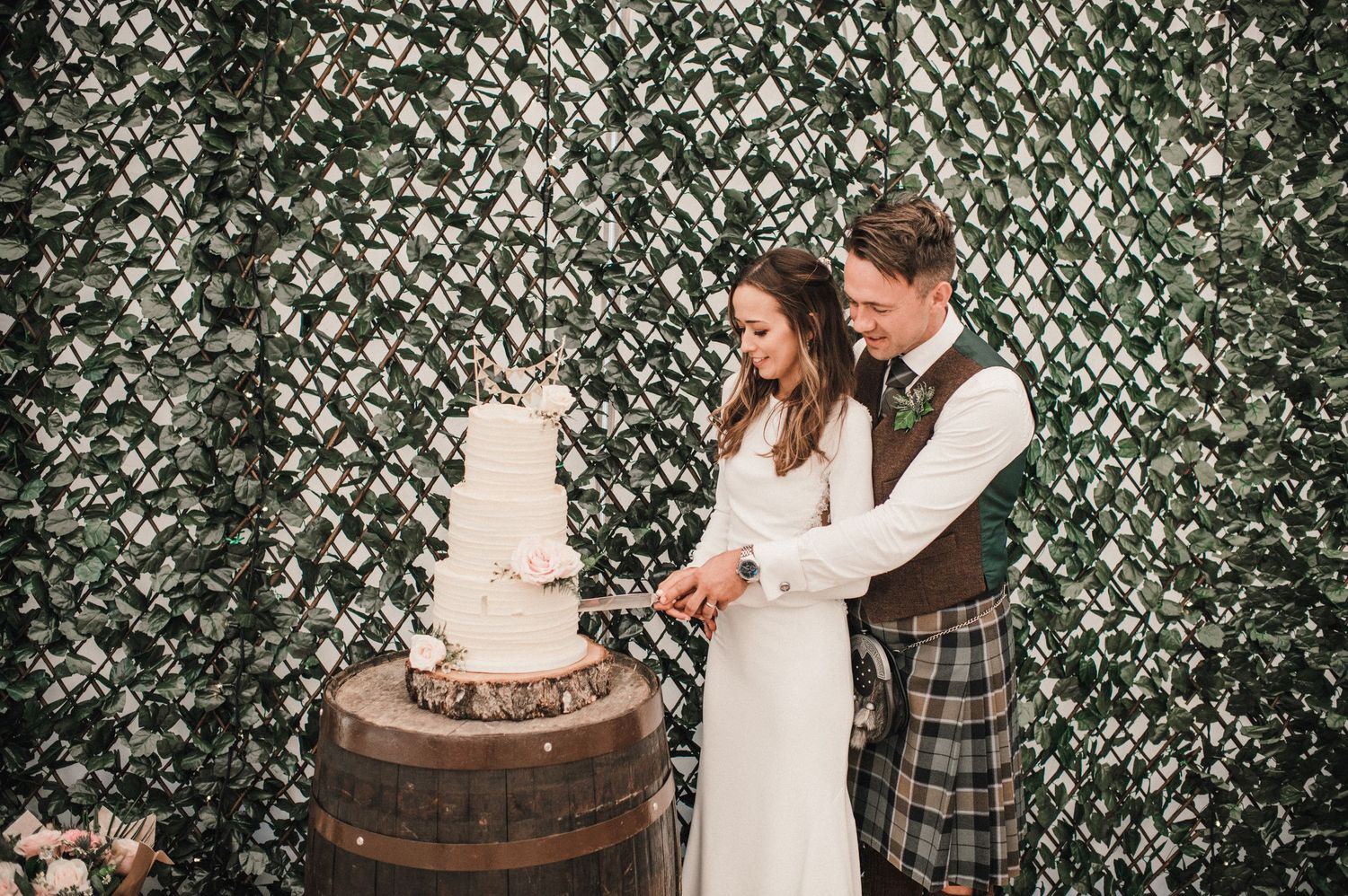 Couple cutting their wedding cake displayed on rustic barrel against ivy backdrop.