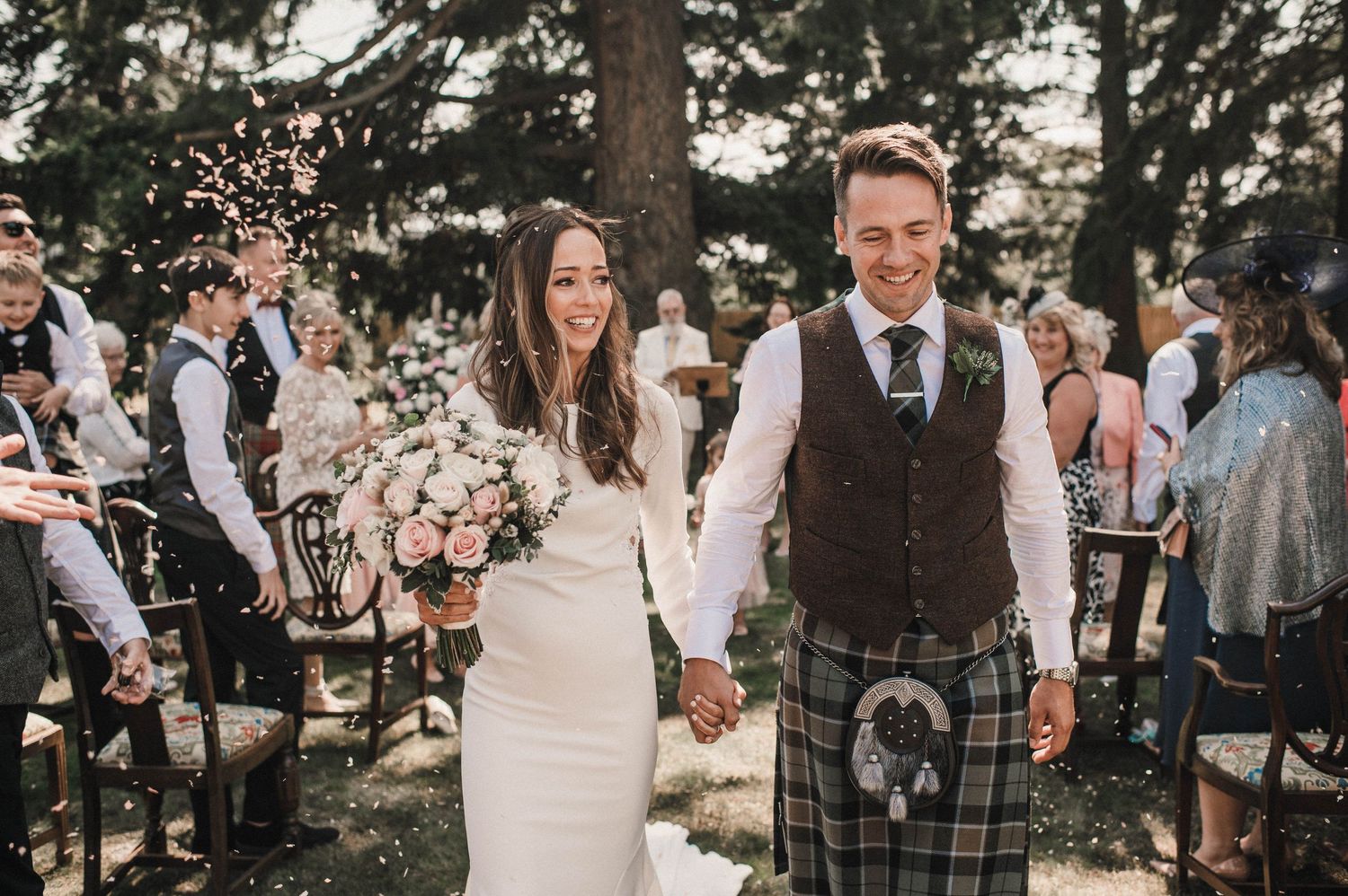 Newlyweds walk hand in hand through their outdoor wedding celebration wearing traditional Scottish attire.