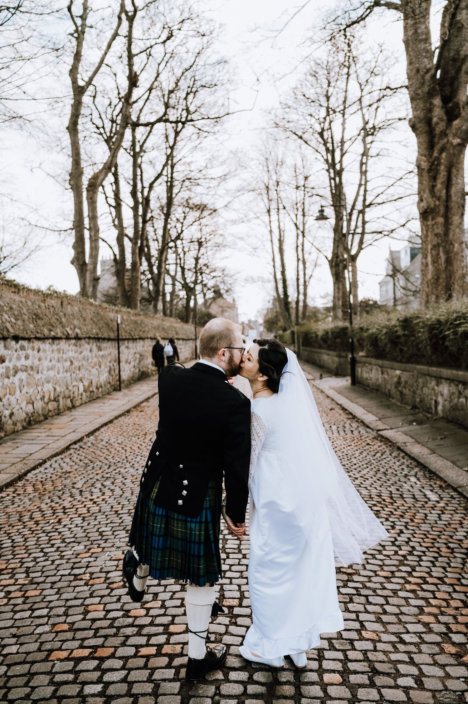 A couple shares an intimate moment while walking on a scenic cobblestone path lined with stone walls.