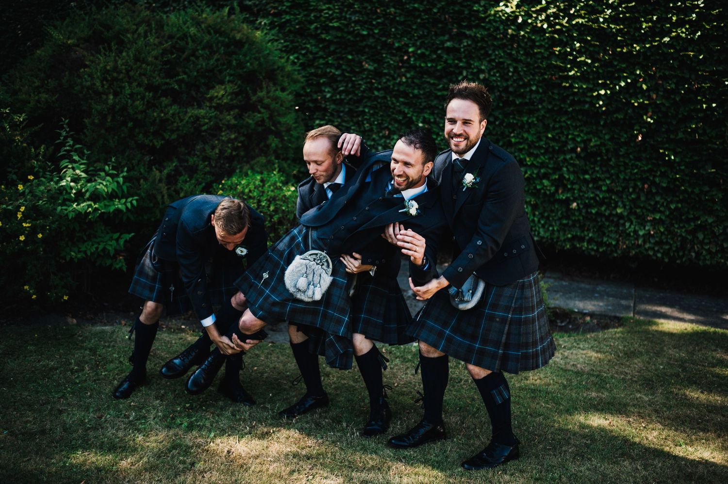 Groomsmen in kilts playfully posing and jumping in garden setting.