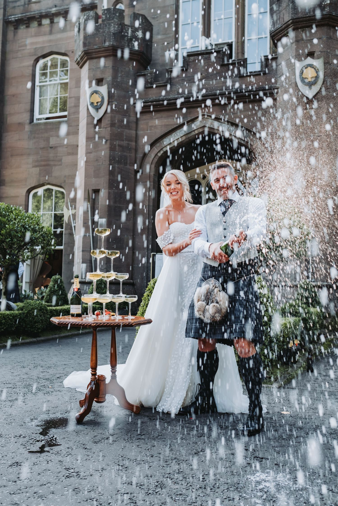 Newlyweds celebrate with champagne spray in front of a castle-like venue with a traditional Scottish kilt and white gown.