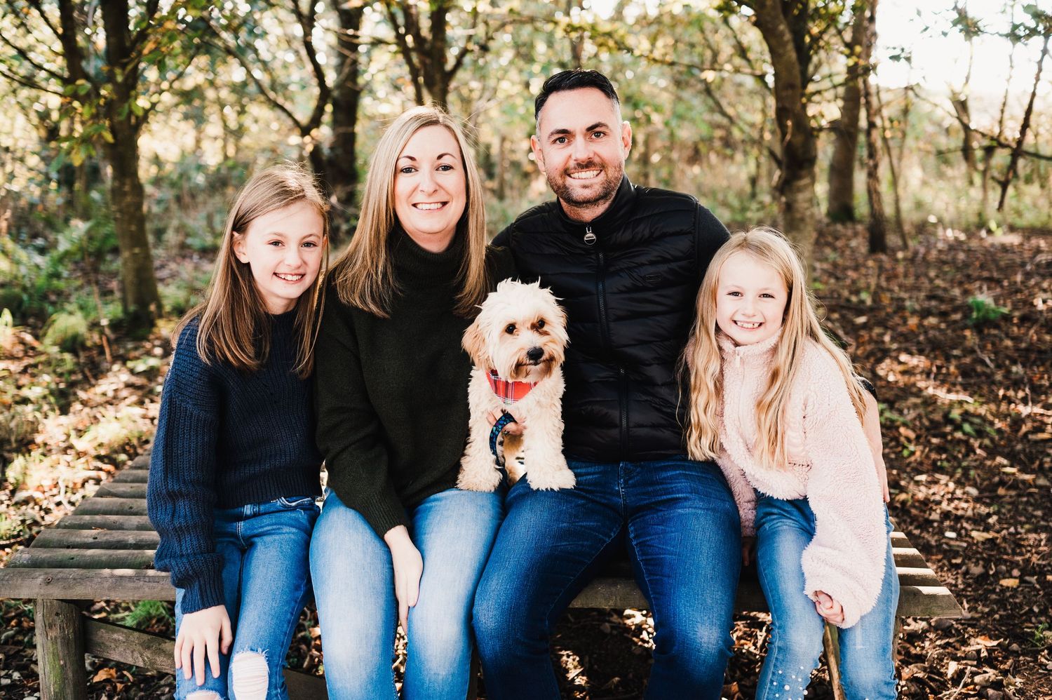 Family sitting on wooden bench in autumn forest with small white fluffy dog wearing orange bow tie.