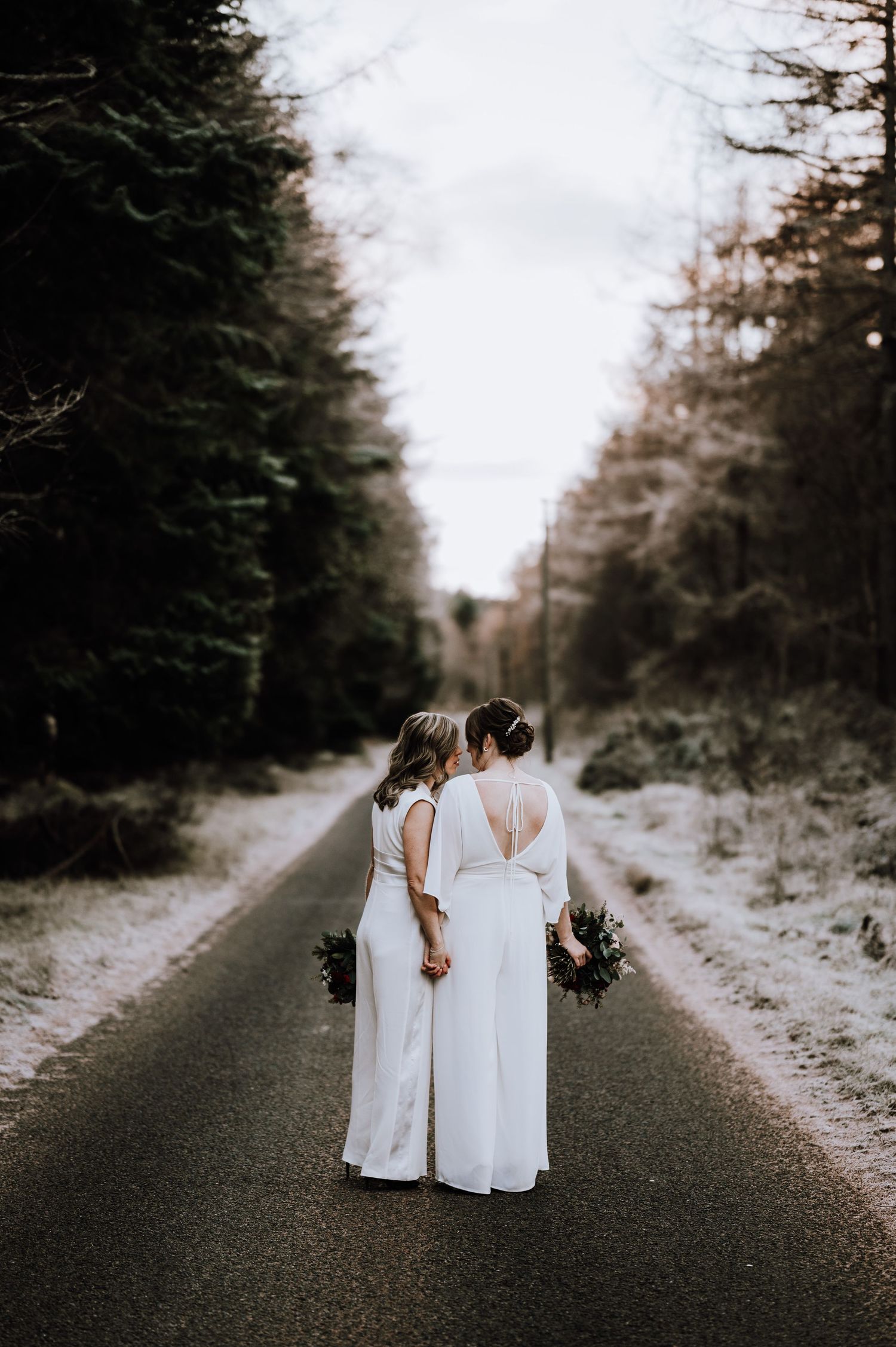 Same Sex Wedding Photography. A couple in matching white dresses walks down a misty forest road holding hands.