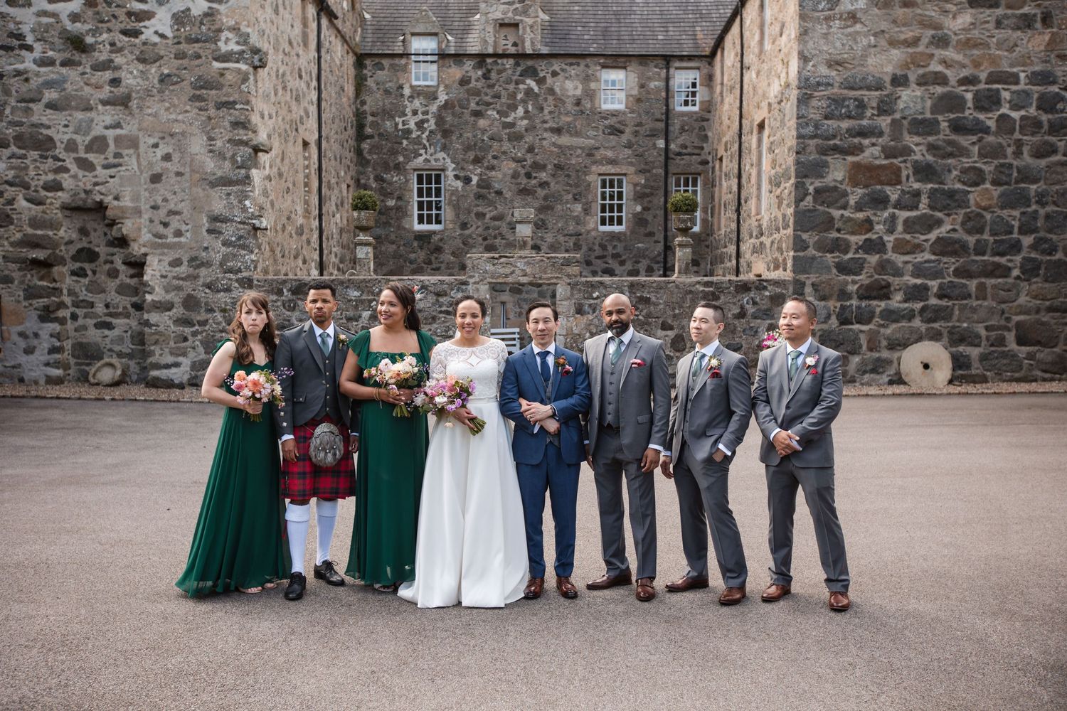 Wedding party poses for formal photos against historic stone building exterior.