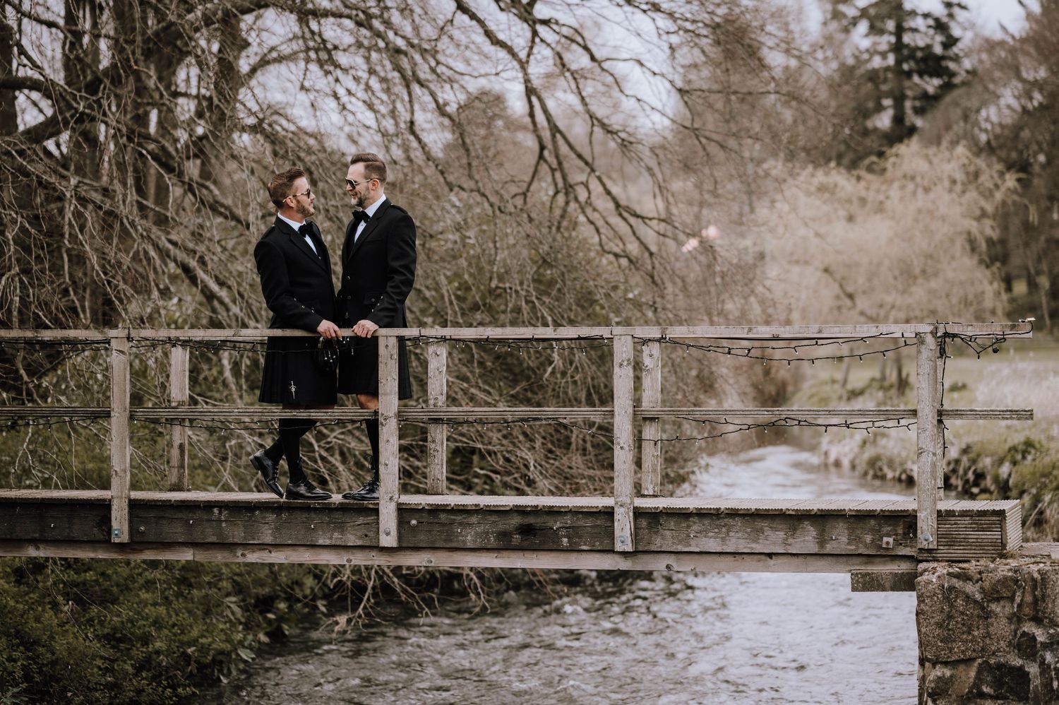 Logie House Wedding Photography. A couple in formal wear stands together on a wooden bridge over water.