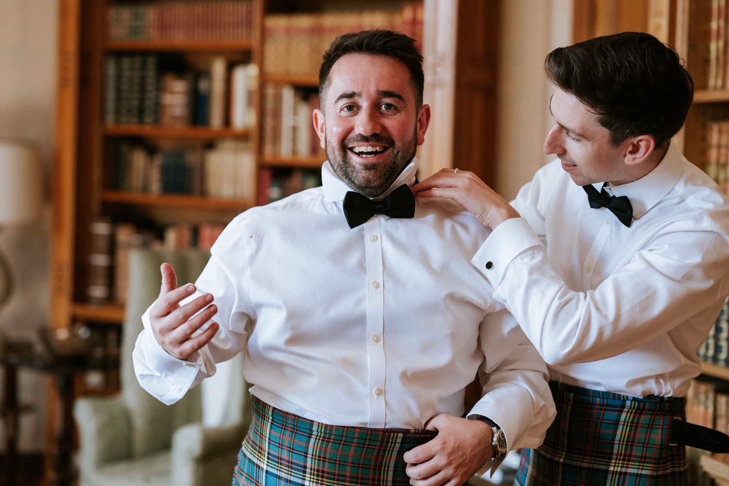 Wedding party members in kilts and formal attire sharing drinks and smiles in an elegant library setting.