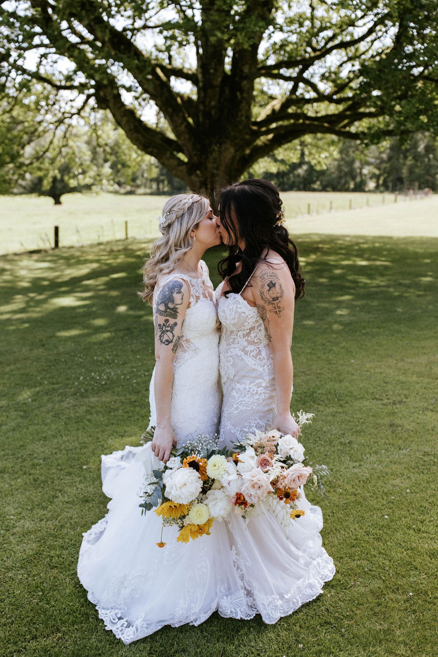 Two brides in matching white dresses holding floral bouquets under a large tree.