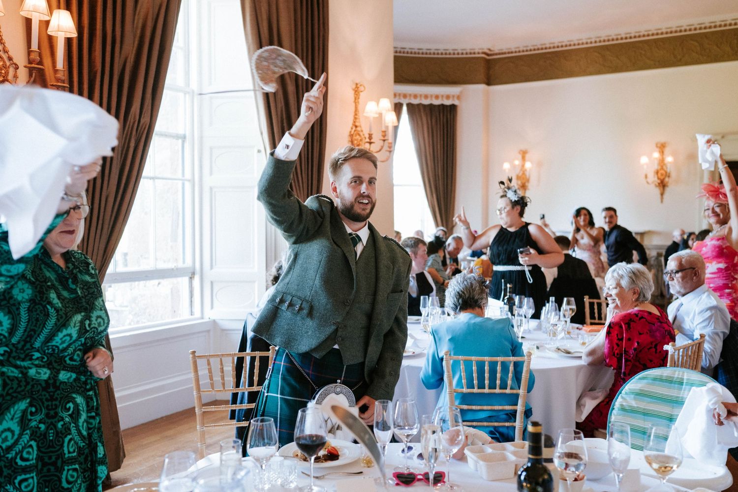 Wedding guests celebrate with raised arms in elegant reception room with chandeliers.