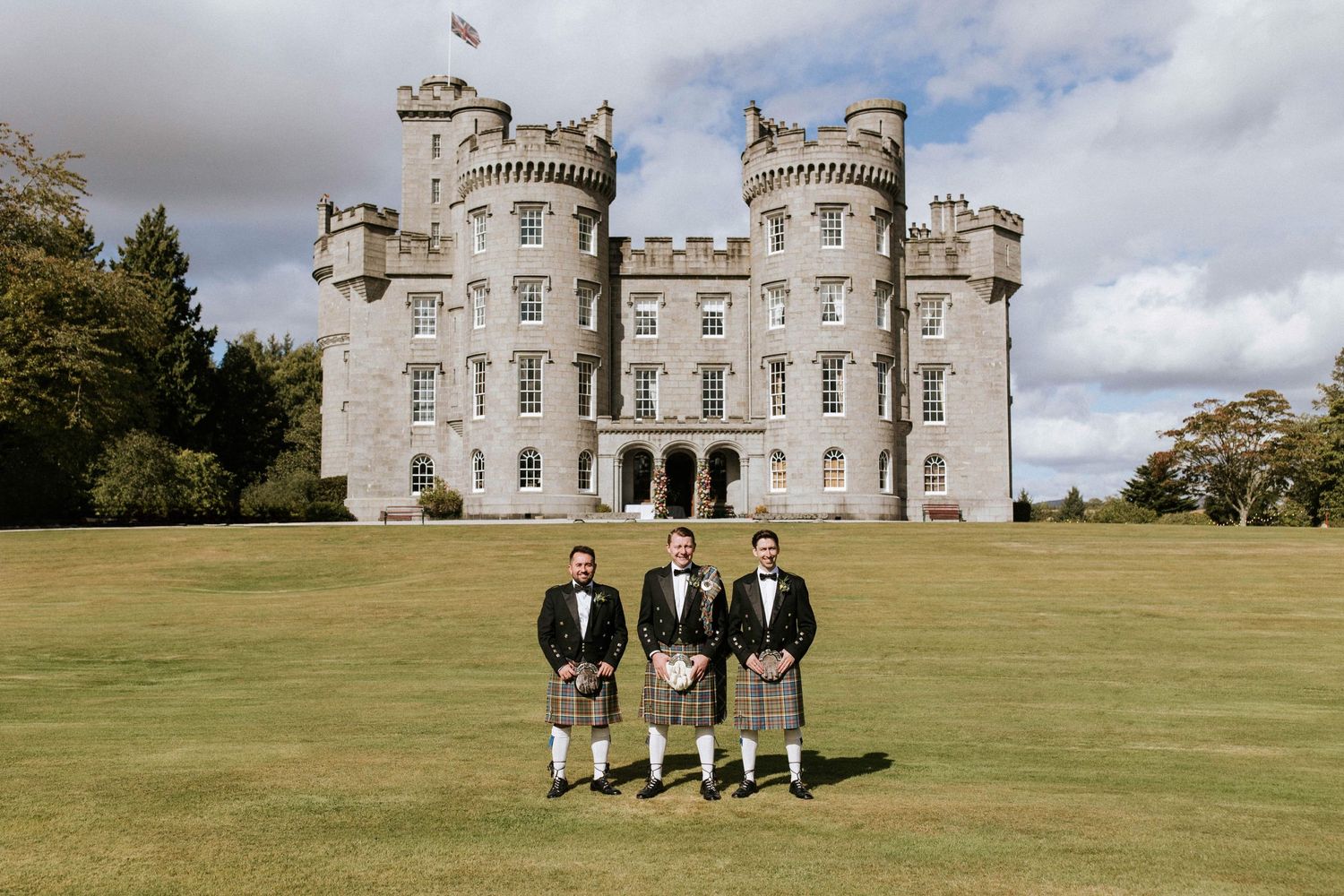 A majestic castle venue with towers sits on a sprawling green lawn under cloudy skies.