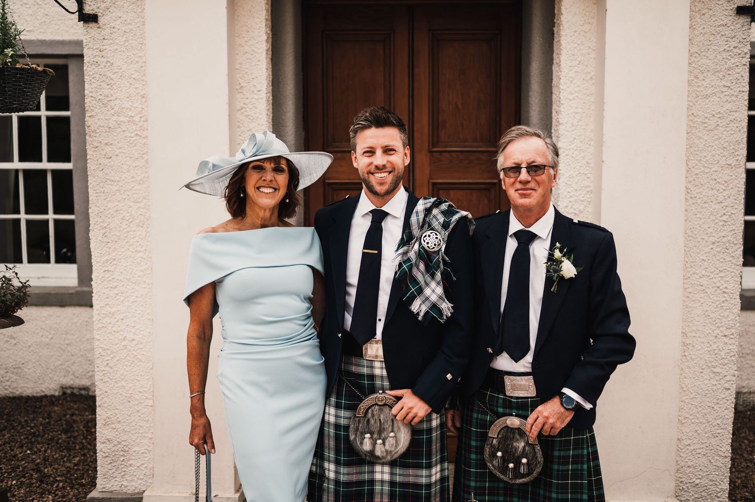 Three people in formal attire pose outside white building entrance for wedding photo.