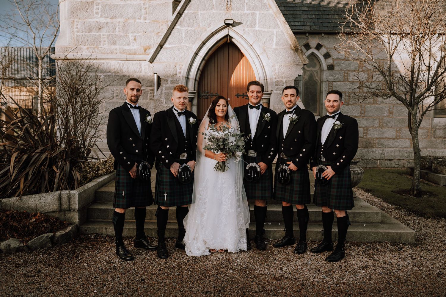 Groomsmen in traditional Scottish kilts and formal wear stand with couple at church entrance.