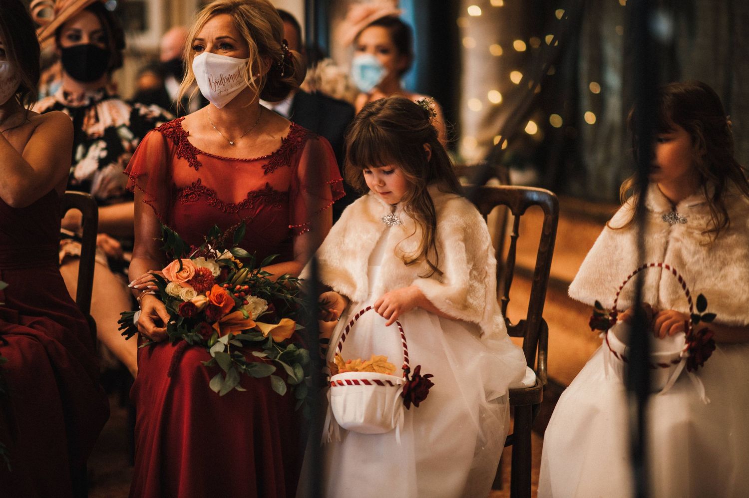 Wedding guests in face masks at an indoor ceremony holding floral bouquets and white lanterns.