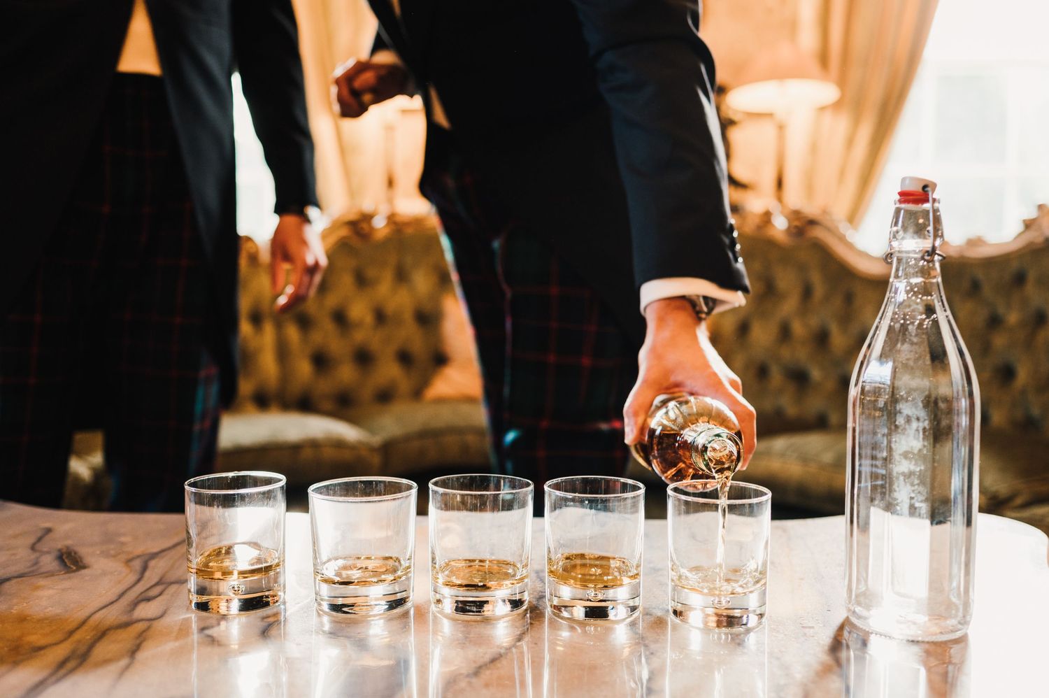 Row of empty shot glasses on a wooden table with someone pouring drinks.
