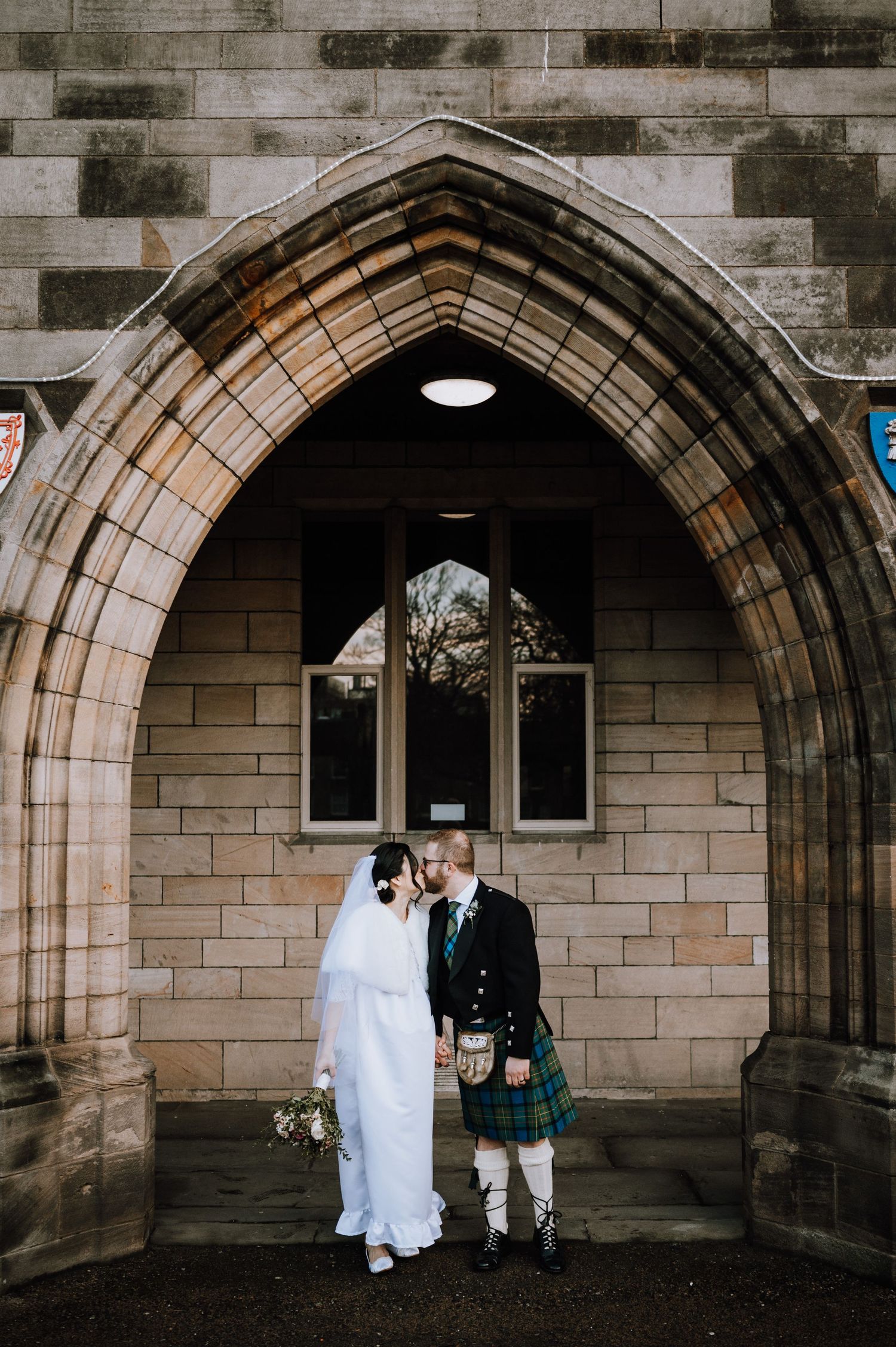 Romantic scene between newlyweds in Scottish and traditional wedding attire against stone wall.