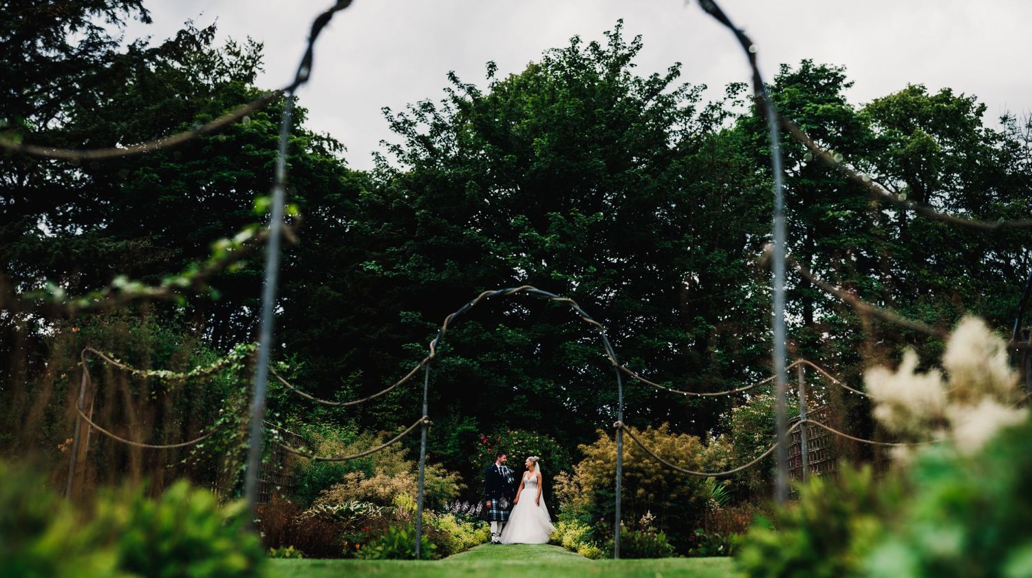 Long distance shot of wedding ceremony setup under rustic garden arch with natural landscaping.