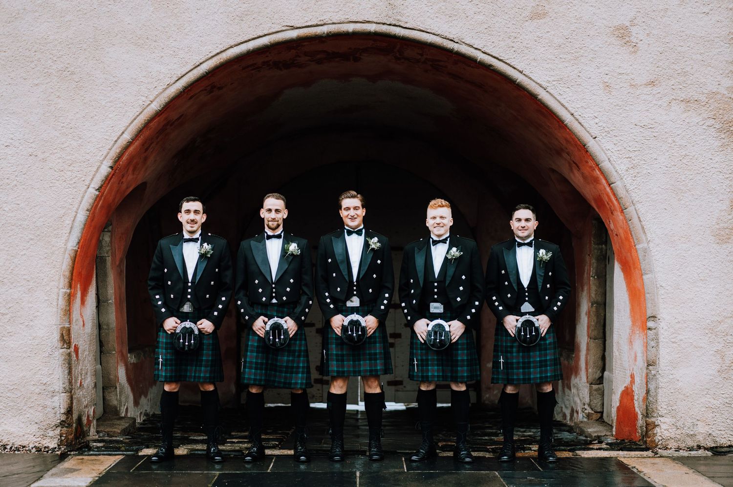 Five groomsmen in matching kilts and formal jackets standing under stone archway.