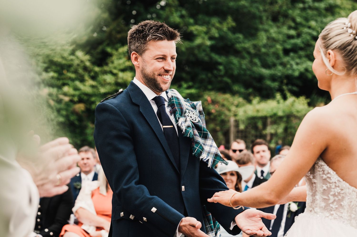 A wedding ceremony in a garden with guests watching as kilted groom exchanges vows with bride.