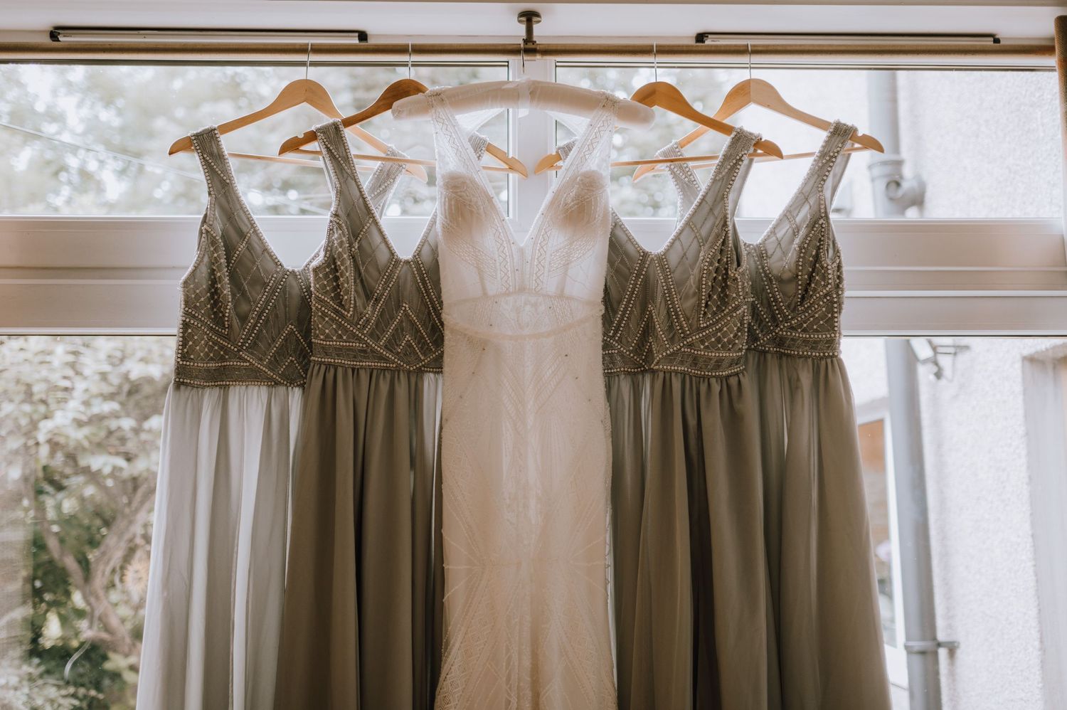 A white wedding gown flanked by sage green bridesmaid dresses hanging by a window.