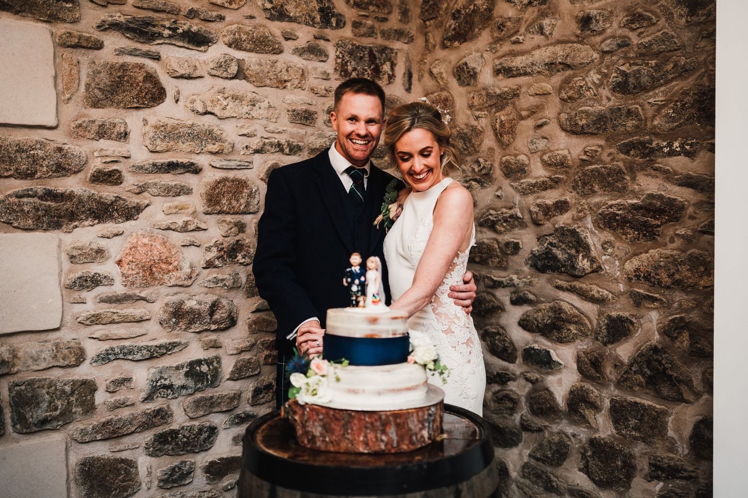 Newlyweds sharing a joyful moment while cutting their navy and white wedding cake in a rustic stone venue.
