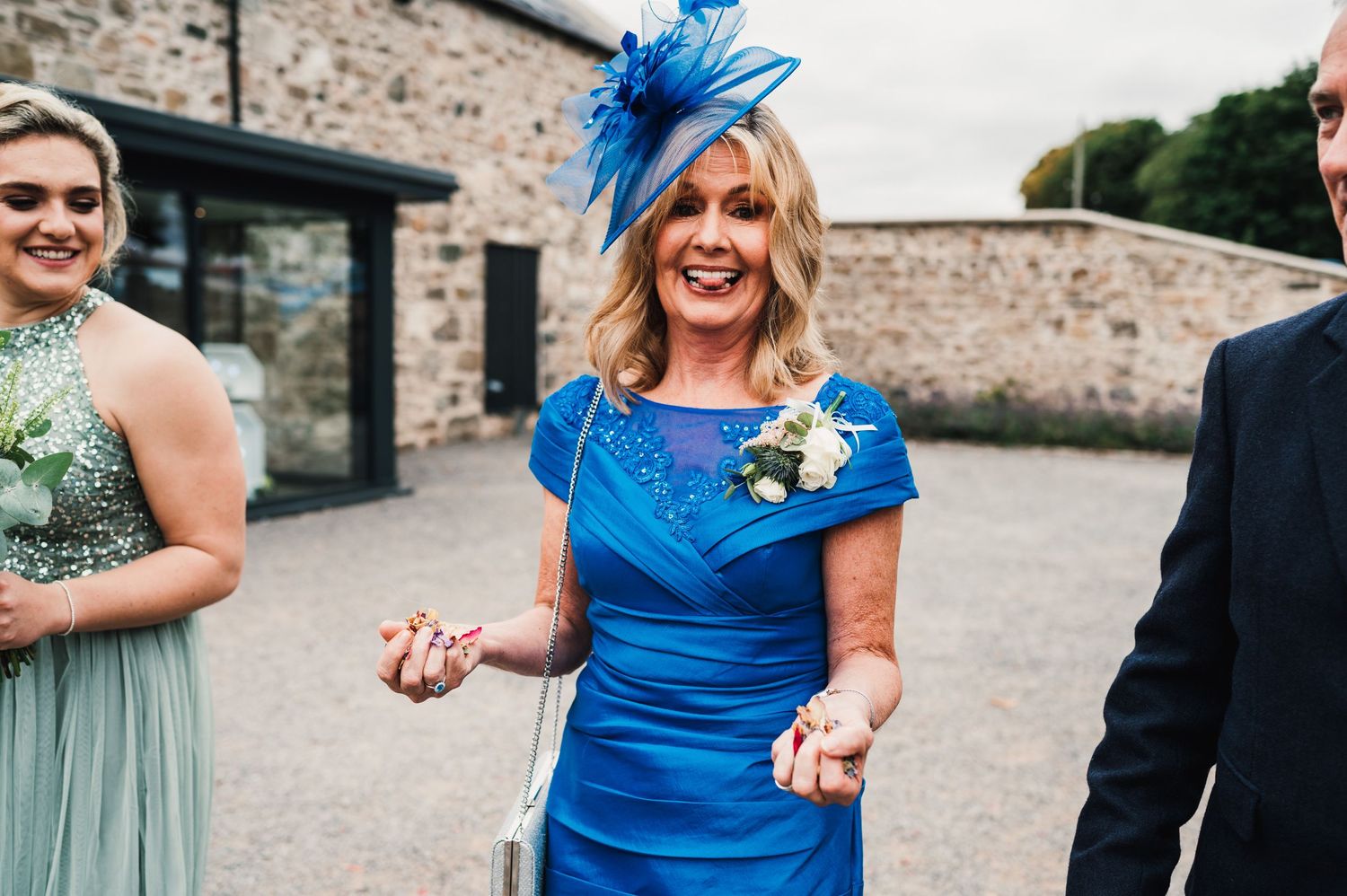 Wedding guest in bright blue dress with matching fascinator smiles joyfully outside stone building.