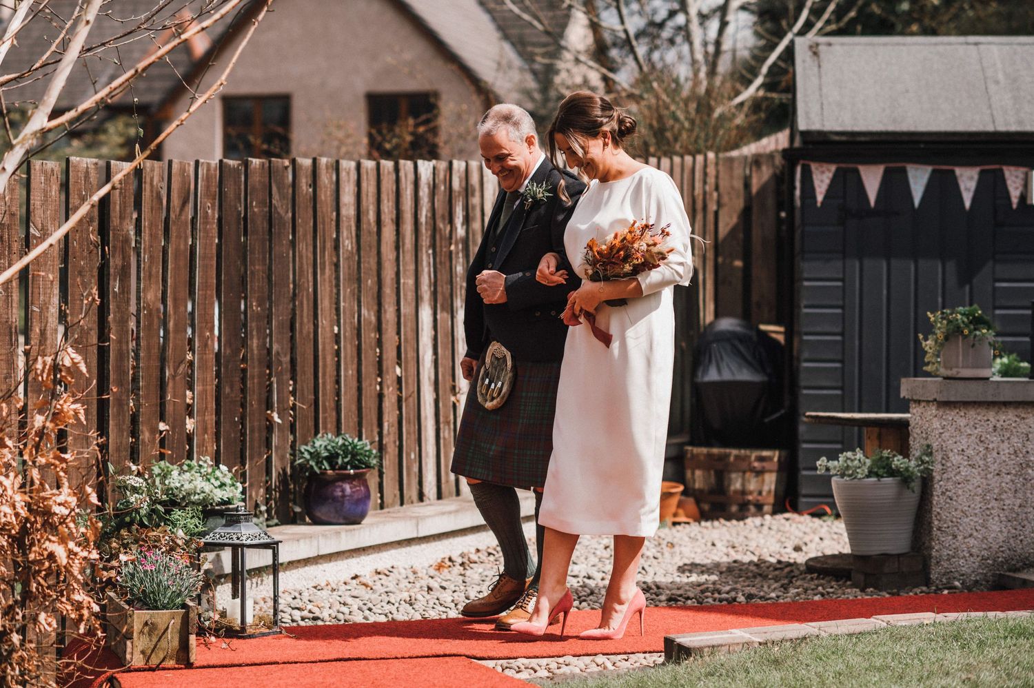 A couple walks along a red carpet path in a garden setting during their Scottish-themed wedding.