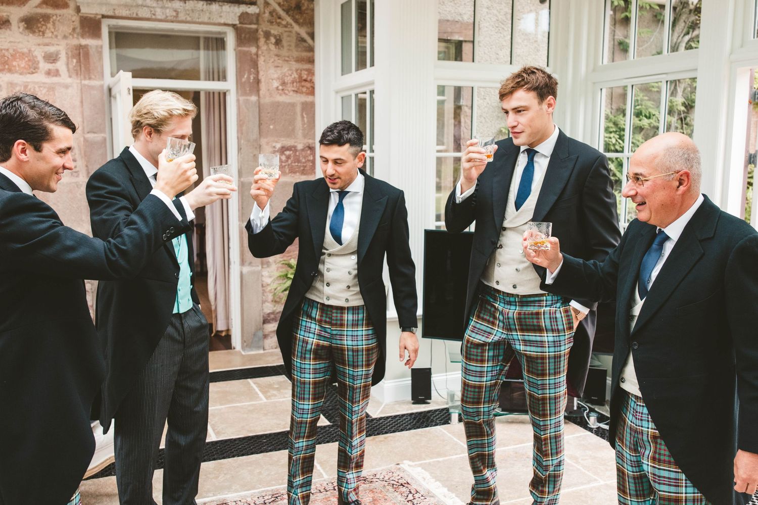 Group of men in formal wear and kilts sharing laughs outside stone building.