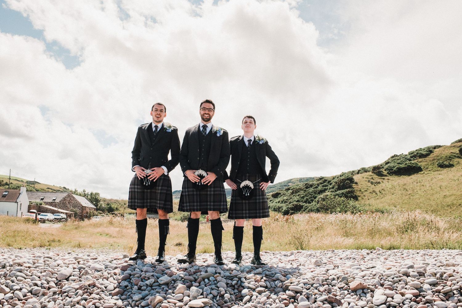 Three groomsmen in traditional Scottish kilts pose together on a rocky beach.
