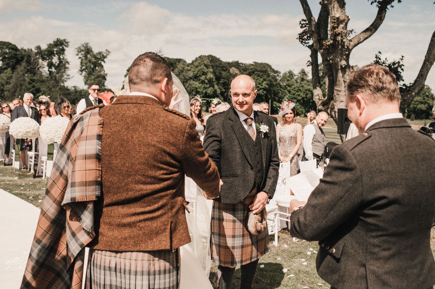 Guests in traditional Scottish kilts and formal wear gather outdoors at the wedding venue.