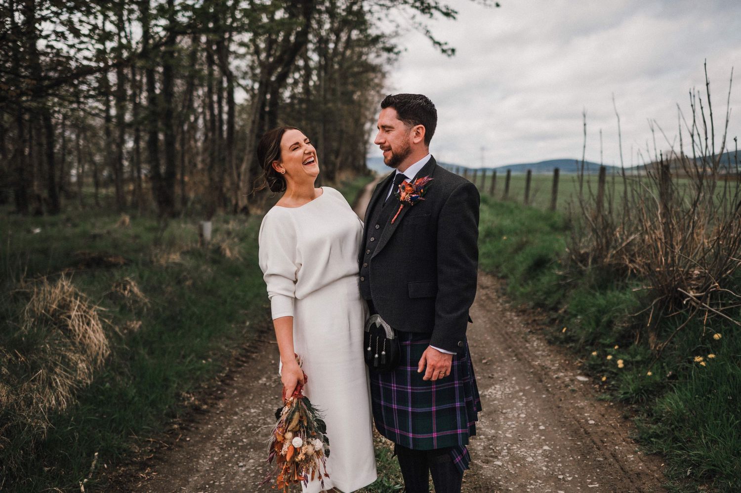 A Scottish wedding couple on a country path with mountains in background, bride in white dress and groom in traditional kilt.