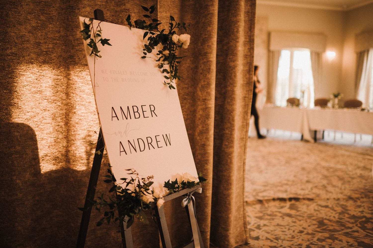 Wedding welcome sign for Amber and Andrew decorated with greenery and illuminated by warm lighting.