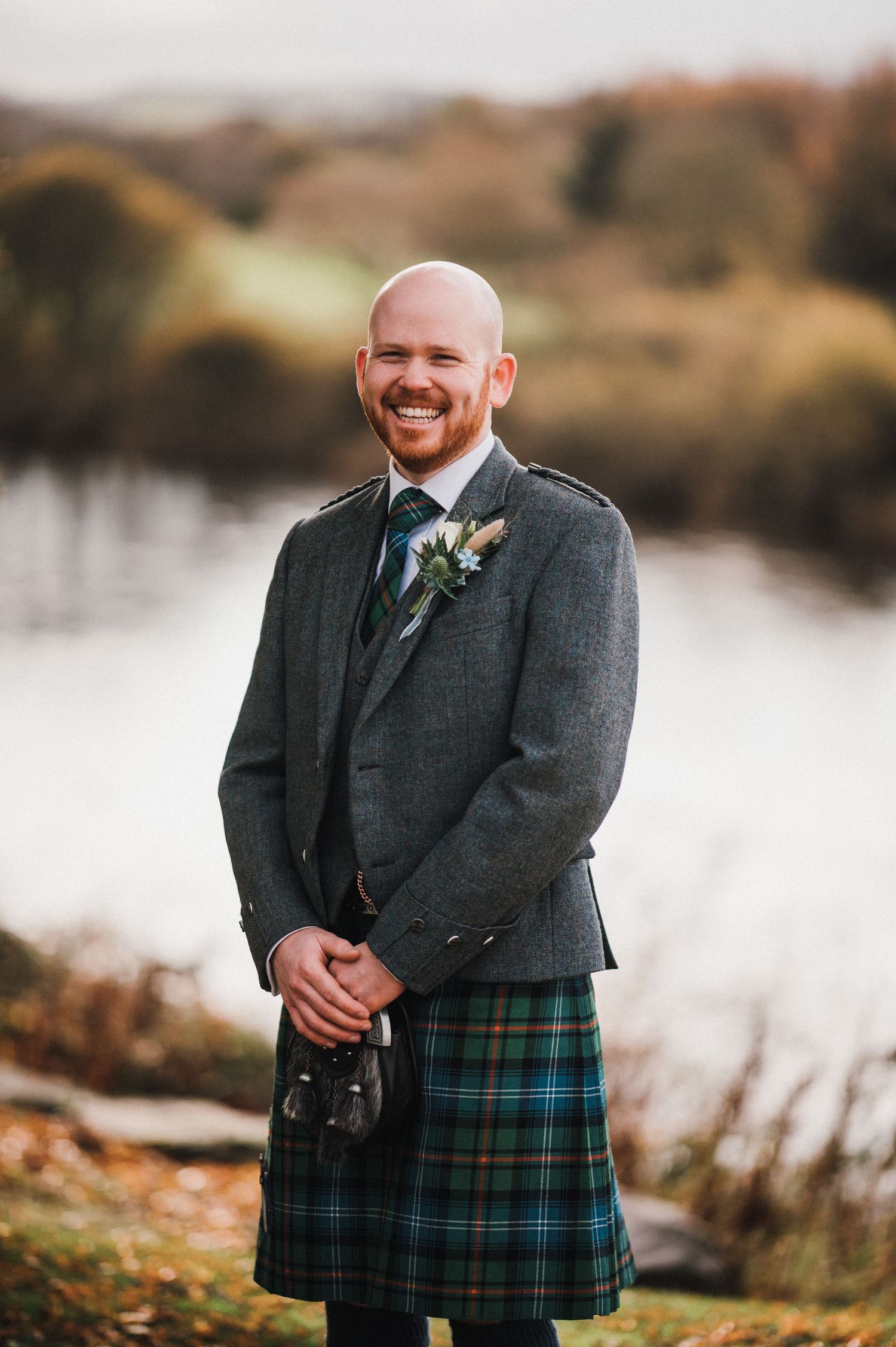 A person in a grey suit jacket and traditional tartan kilt poses by a lake during autumn.
