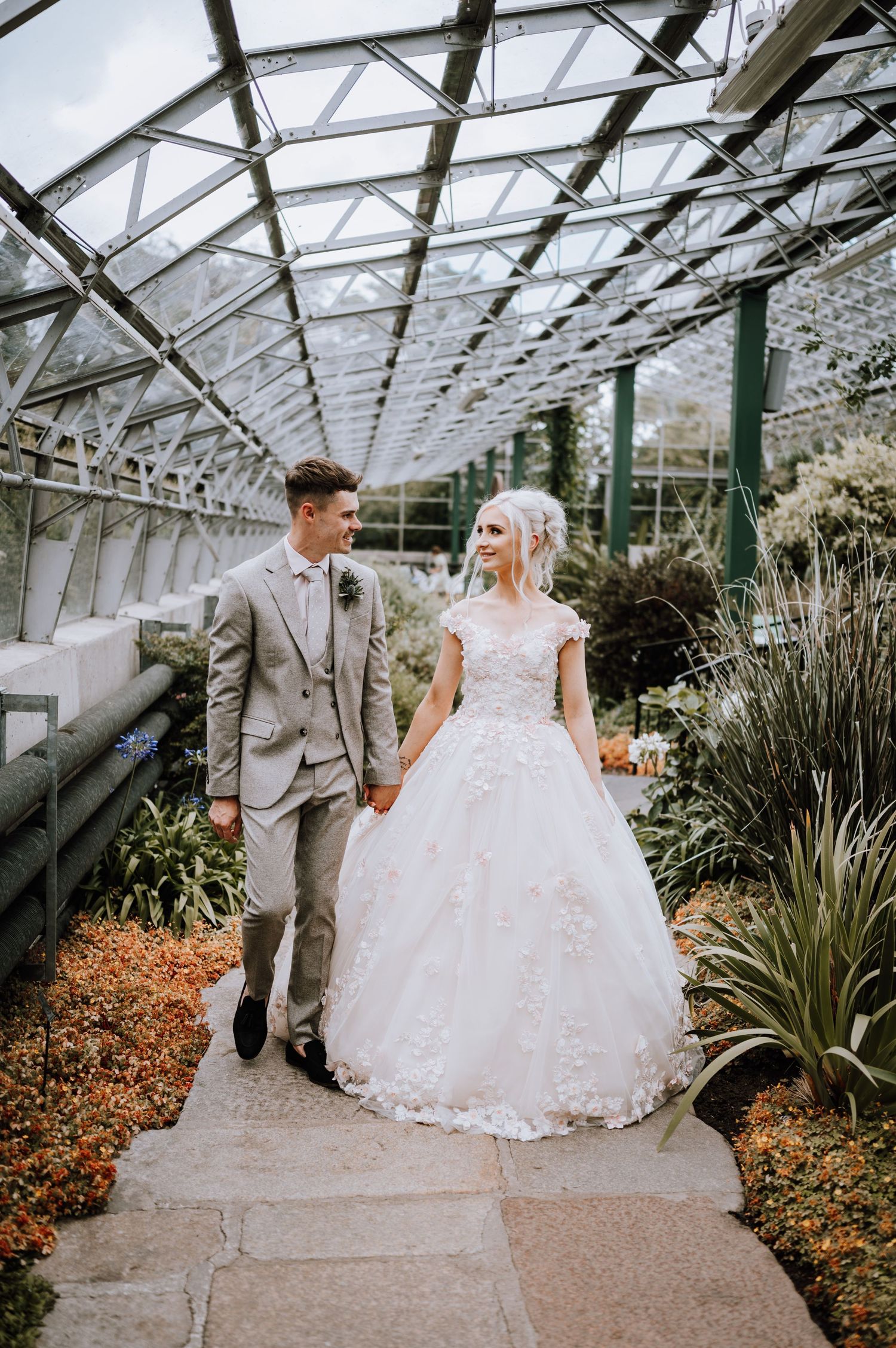 A wedding couple walks hand in hand through a glass-roofed conservatory garden.
