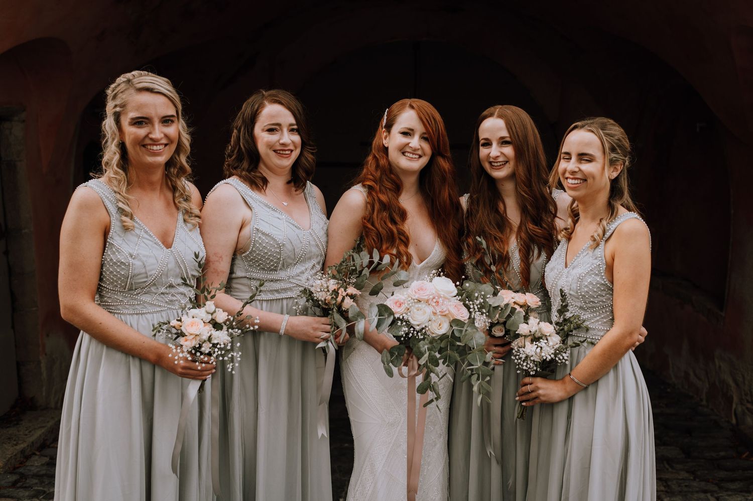 Five women in grey dresses holding bouquets of pink and white flowers with greenery stand together.