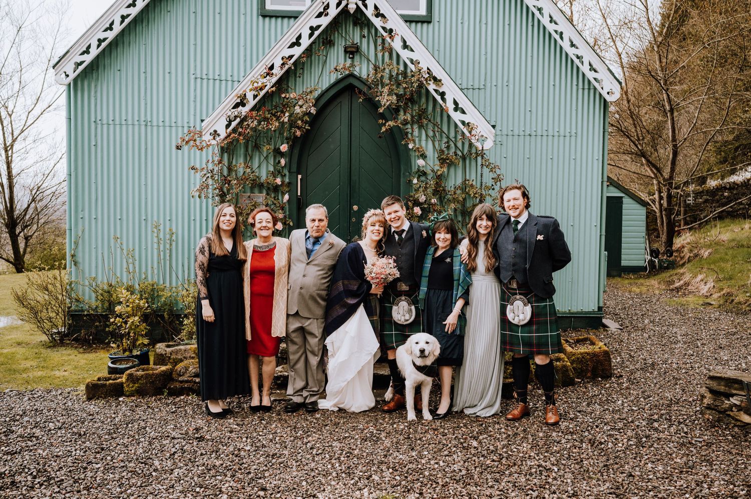 Large wedding party with dog poses in front of mint green chapel.