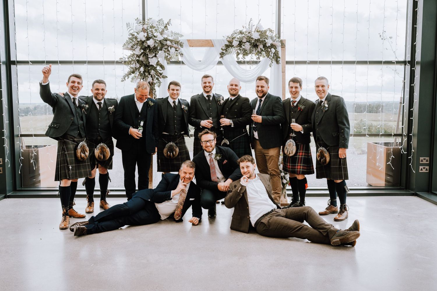 Group of groomsmen in kilts and suits pose together at wedding venue.