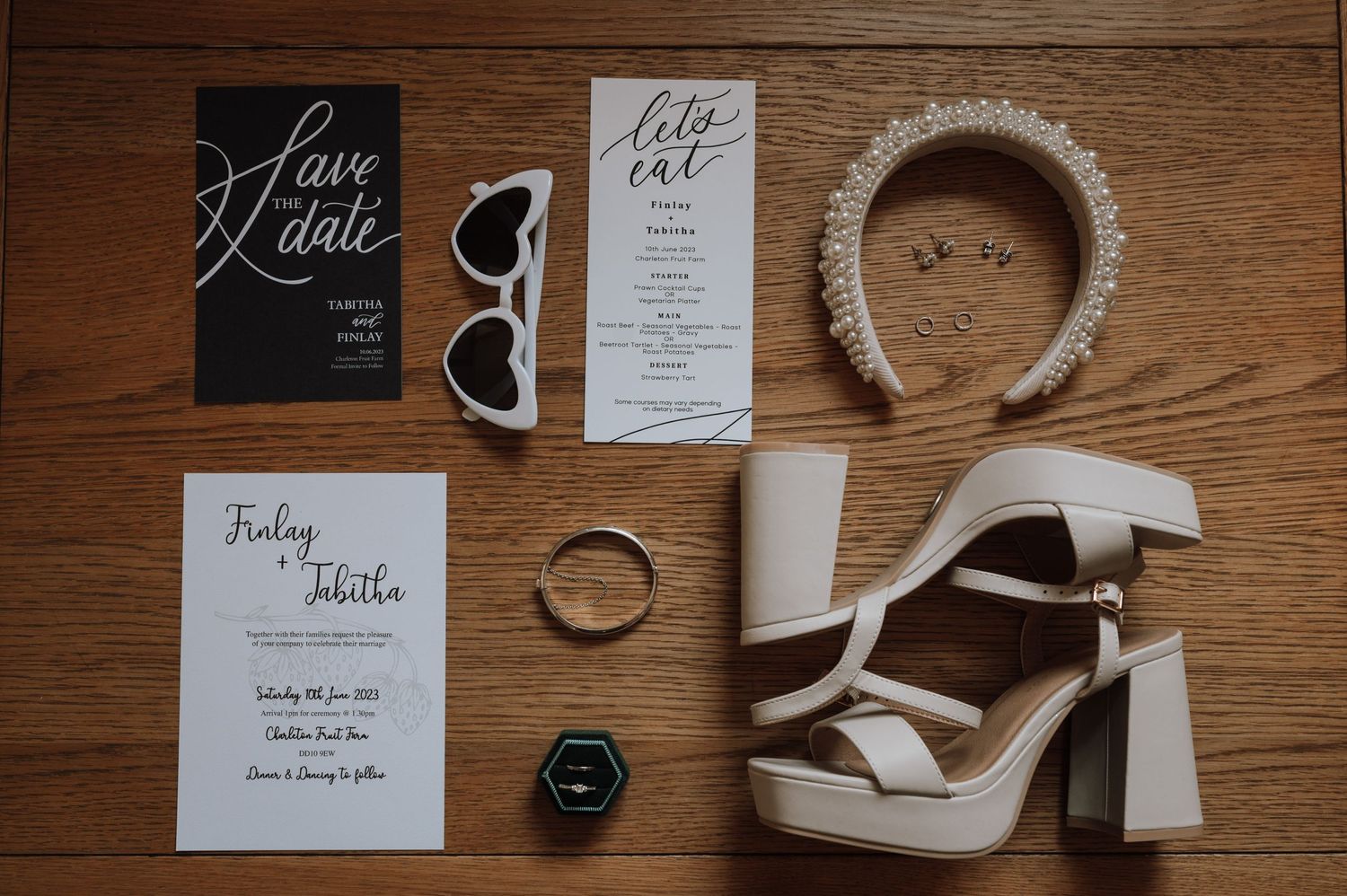 Wedding details flatlay with invitations, sunglasses, headband, and white platform sandals on wooden floor.
