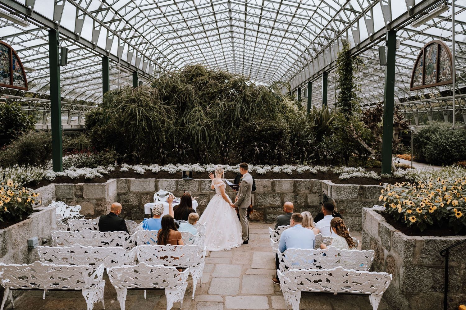 Wedding ceremony setup under a glass greenhouse structure with white chairs and stone walls.