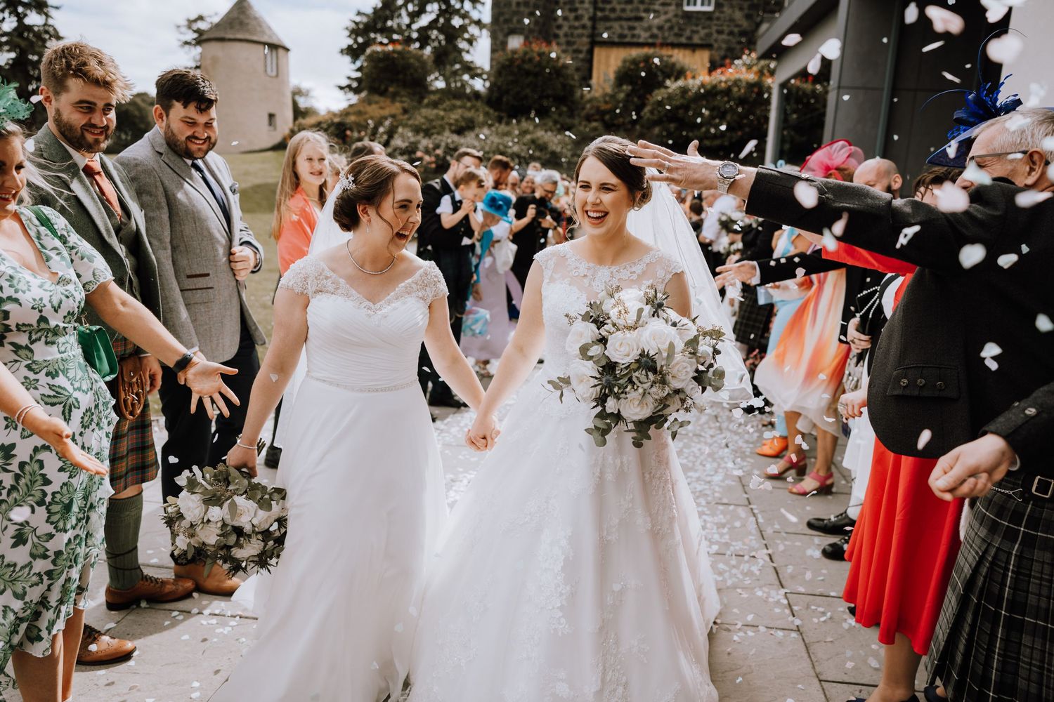 A celebratory wedding exit with confetti as two brides walk hand in hand past guests in Scottish attire.