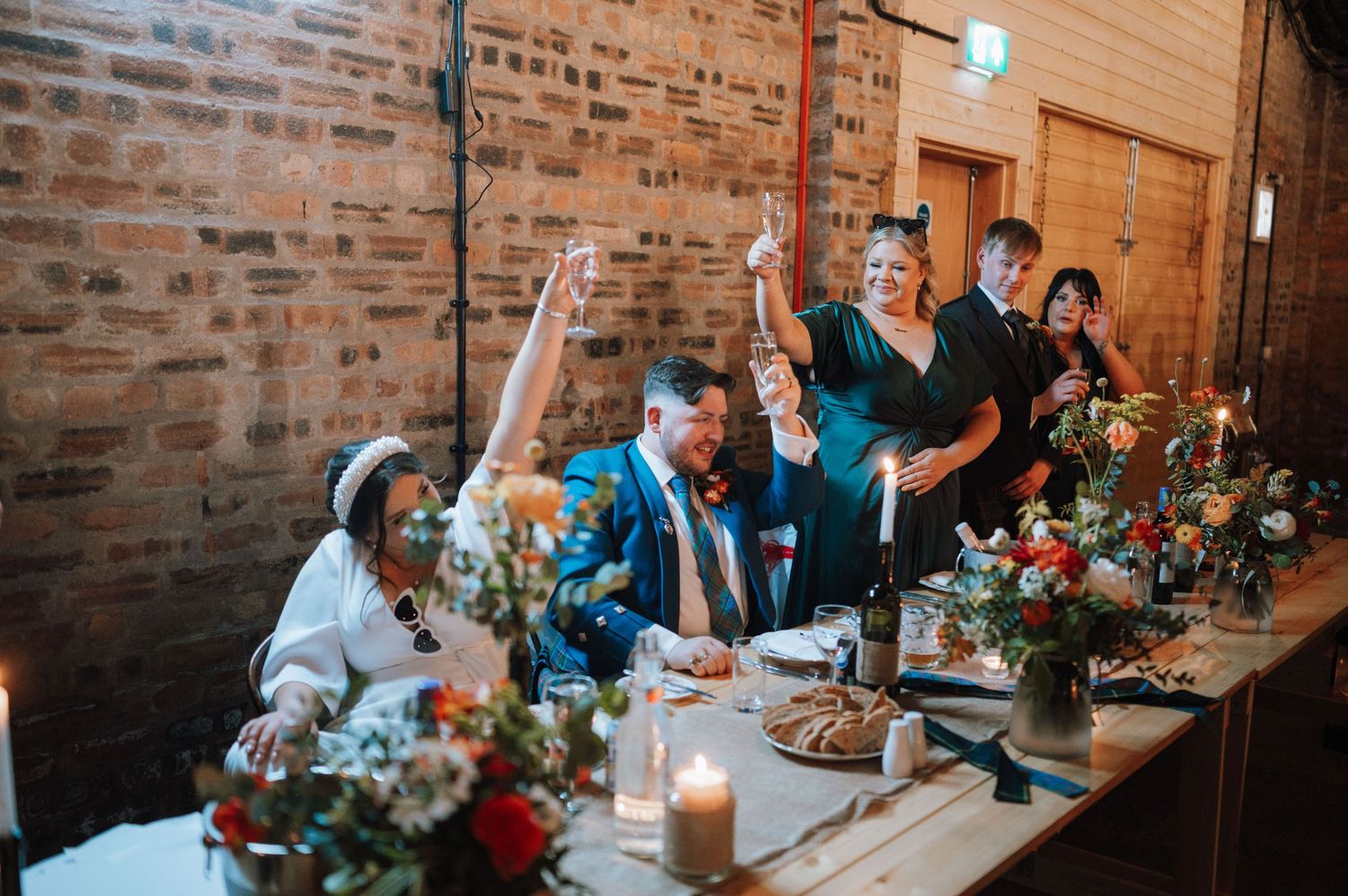 Wedding guests celebrate enthusiastically at a long dining table with floral centerpieces and candlelight.