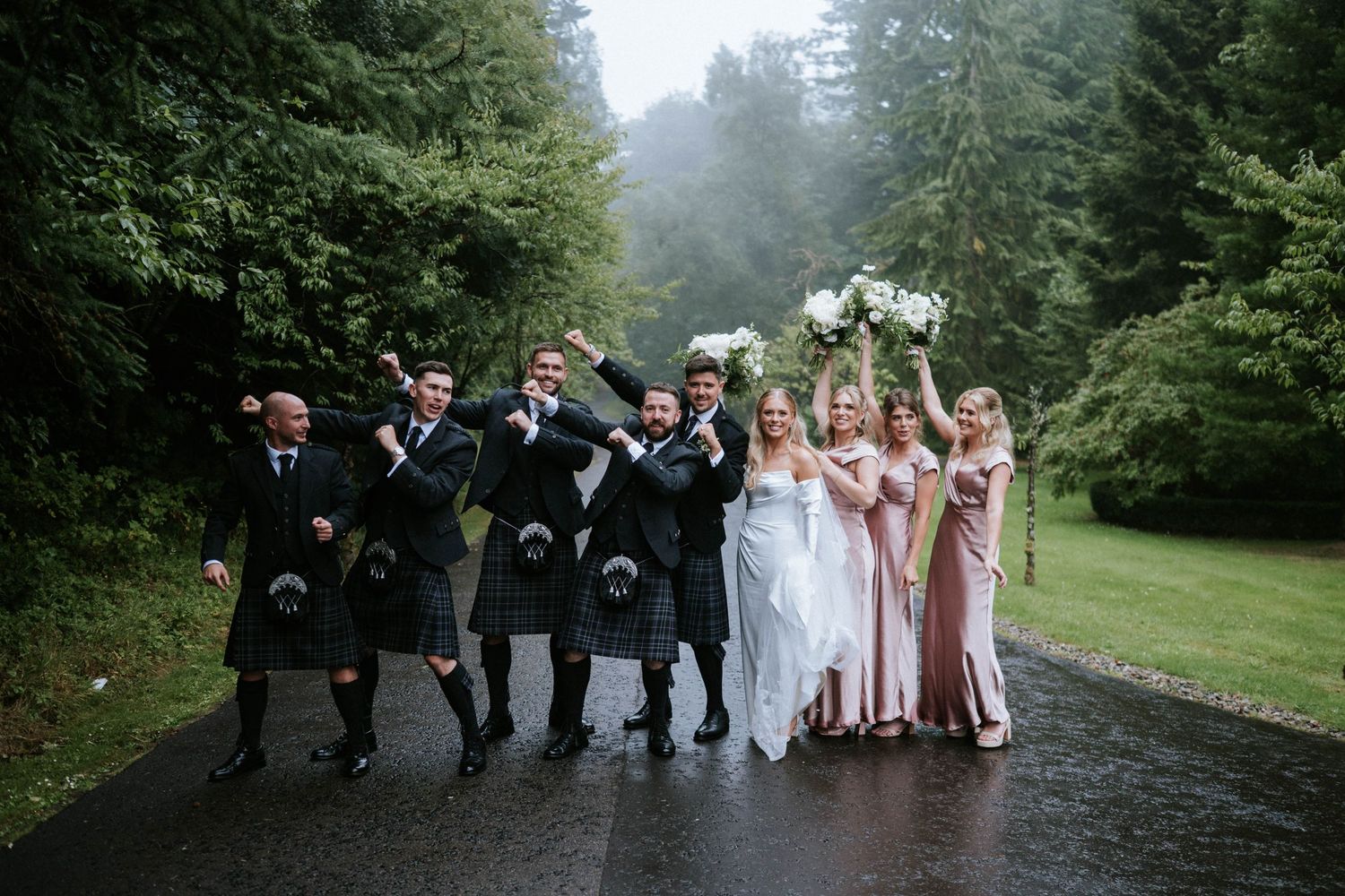 Wedding party walking along misty garden path in formal Scottish attire.