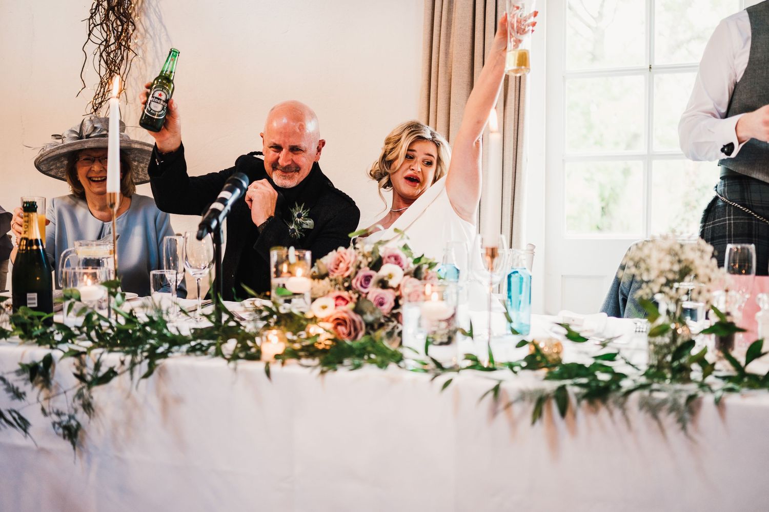 Wedding reception guests celebrate at decorated banquet tables with floral centerpieces and string lights.