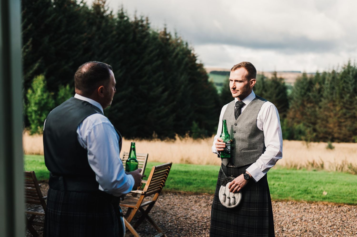 Two servers in formal attire stand outdoors near evergreen trees with one holding drinks at a Scottish wedding.