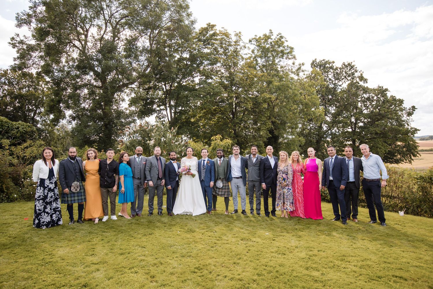 Large wedding party group photo on a green lawn with trees in background.