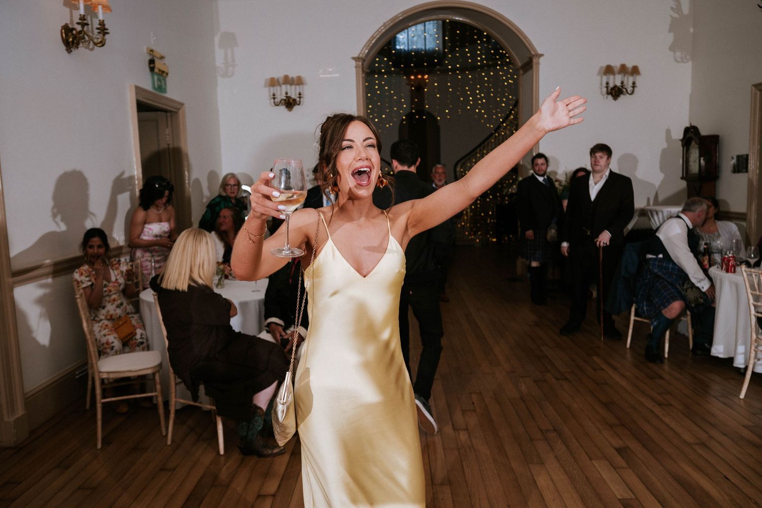 Fasque Castle Wedding Photography. Wedding guests dance and celebrate in a bright ballroom with arched doorways and wall sconces.