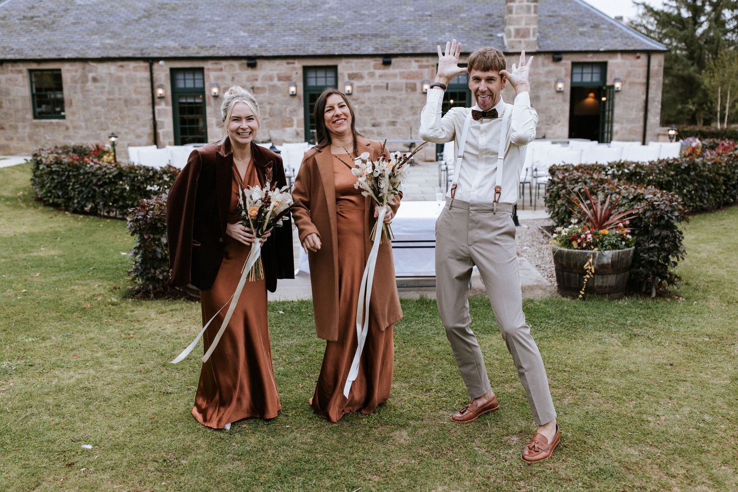 Wedding party members in earth-toned attire posing on lawn in front of stone building.