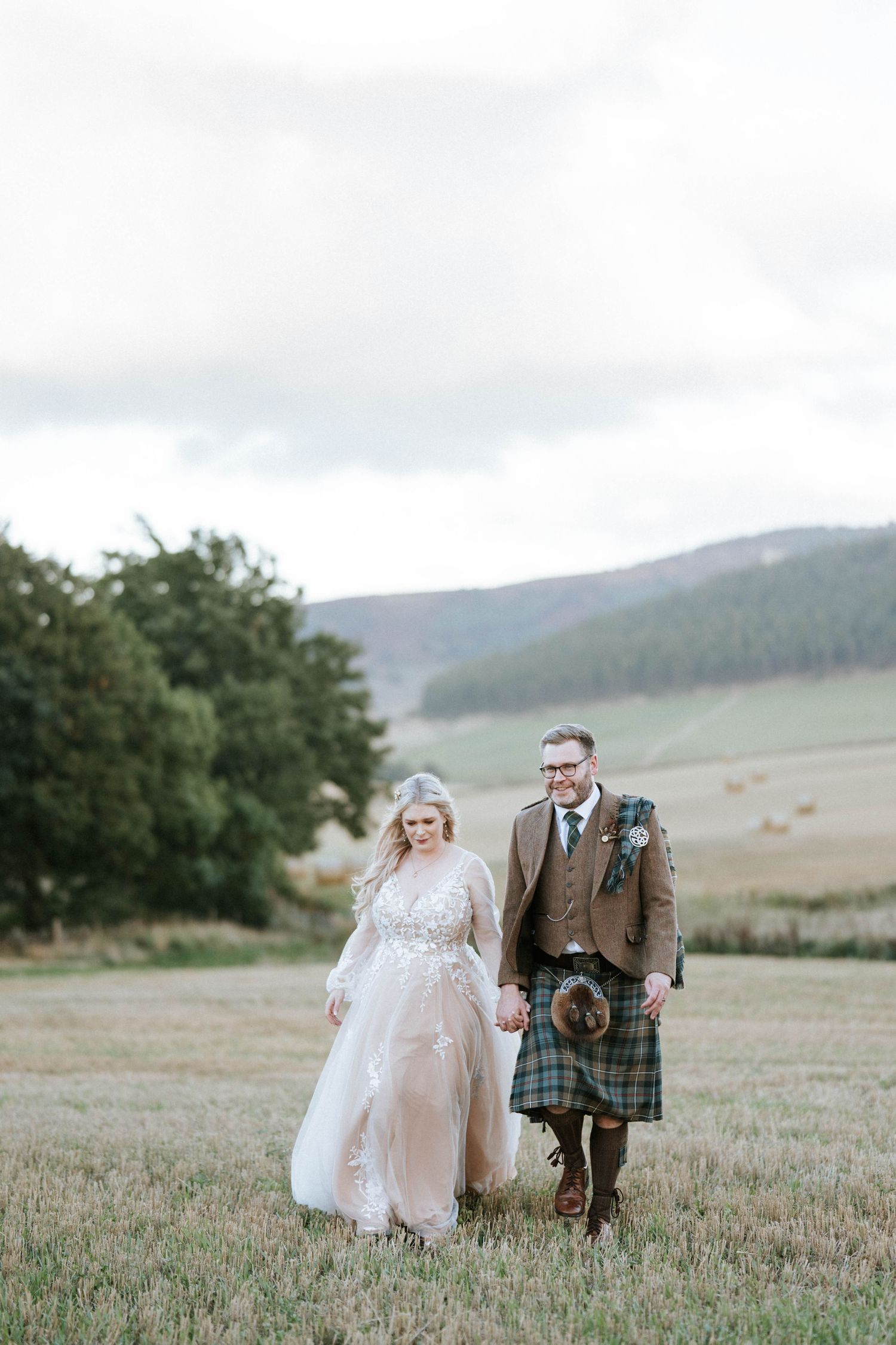 Couple walking hand in hand across a scenic Scottish highland field in wedding attire.