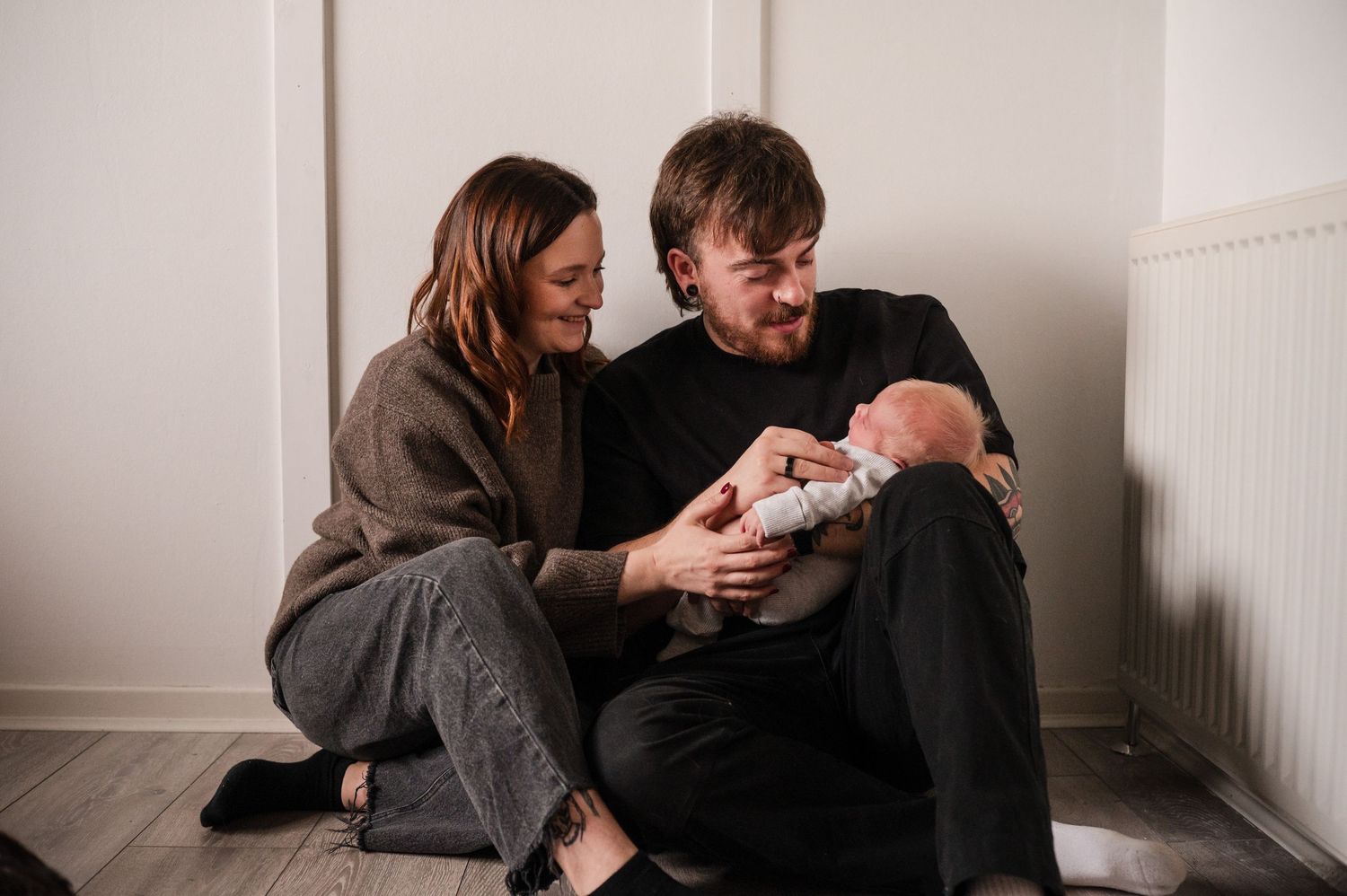 Parents in brown sweaters share an intimate moment with their newborn baby in a minimalist setting.