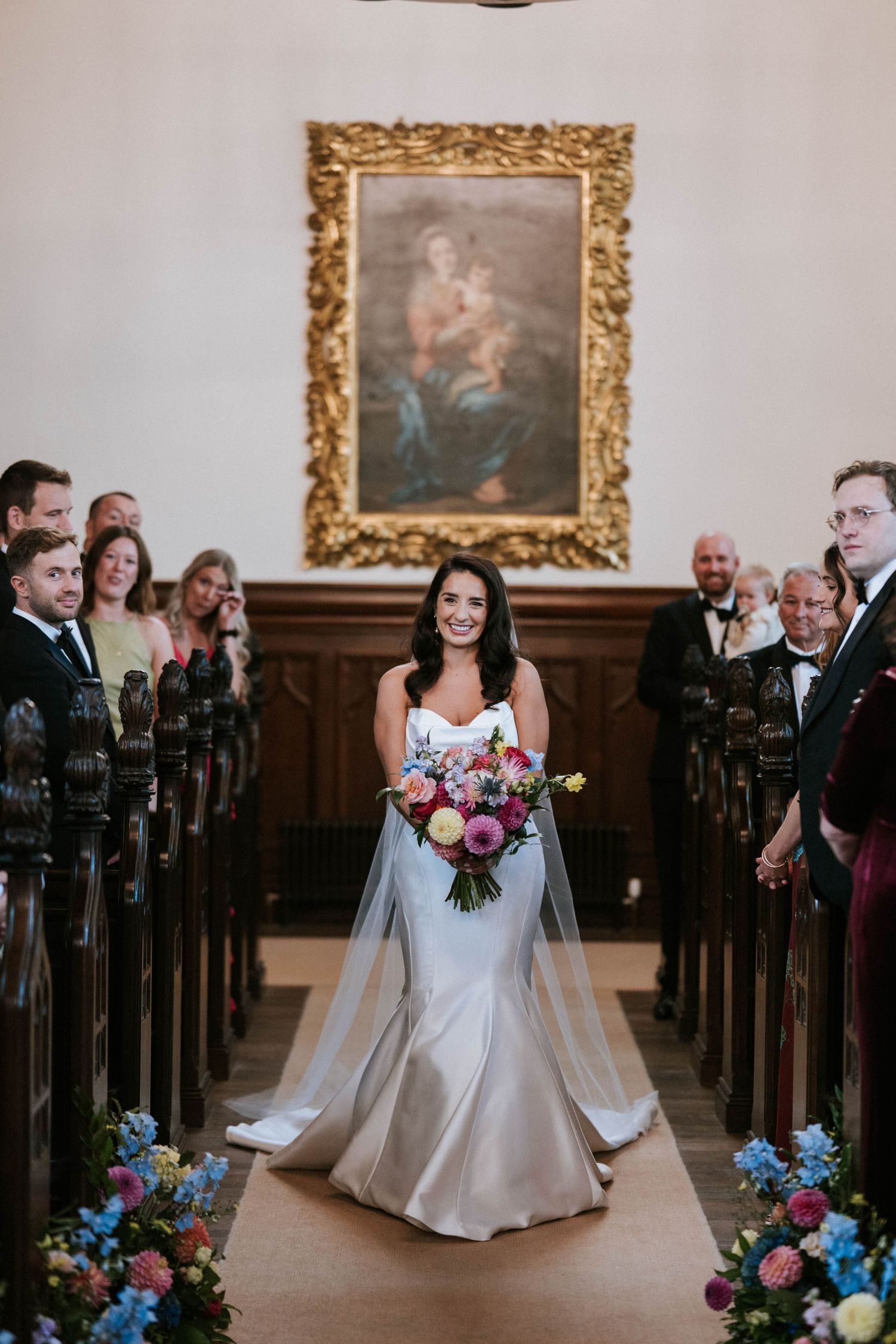 A bride walks down the church aisle decorated with colorful floral arrangements and gold-framed artwork.