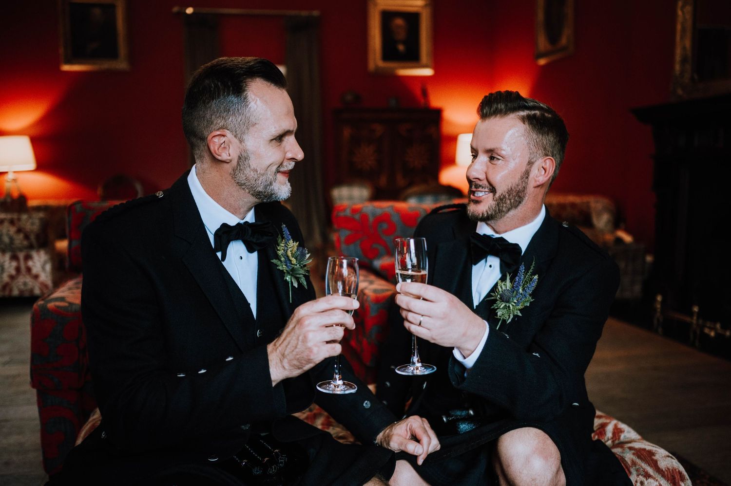 Logie House Wedding Photography. Two people in black tuxedos share drinks in a warmly lit reception venue.